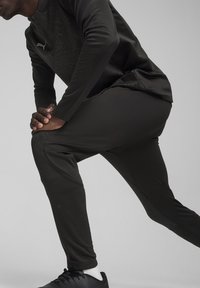 Sportoutfit met een nauwsluitend longsleeve shirt met een patroon op de borst en nauwsluitende broek, beide van donker zwart materiaal, en zwarte schoenen.