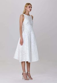 Femme portant une robe midi sans manches blanche à motifs floraux et des sandales à talons blancs, se tenant devant un fond clair.