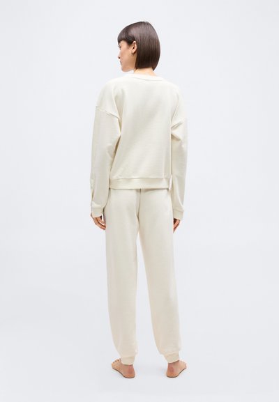 Γυναίκα με κοντά μαλλιά που στέκεται ξυπόλυτη, φορώντας μια φαρδιά, ανοιχτόχρωμη, κρεμ φούτερ μπλούζα και ταιριαστό παντελόνι φόρμας, με την πλάτη προς τα εμπρός.