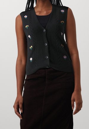 Femme portant un cardigan noir sans manches boutonné avec de petites fleurs brodées violettes et jaunes sur un haut noir et une jupe foncée.