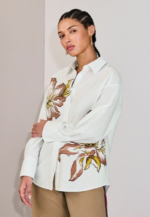 Mujer con cabello trenzado, vestida con una camisa blanca de botones con grandes estampados florales marrones y amarillos, y pantalones beige, sobre un fondo liso.