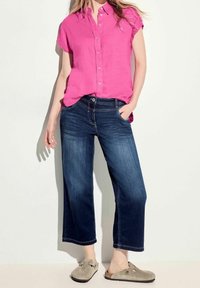 Camicetta rosa a maniche corte con bottoni abbinata a jeans a gamba larga blu scuro e scarpe slip-on grigie. Tessuto morbido con vestibilità comoda.