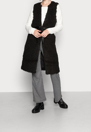 Femme portant un long gilet noir duveteux avec une ceinture nouée, un pull blanc côtelé, un pantalon à carreaux noir et blanc, et des bottines noires.