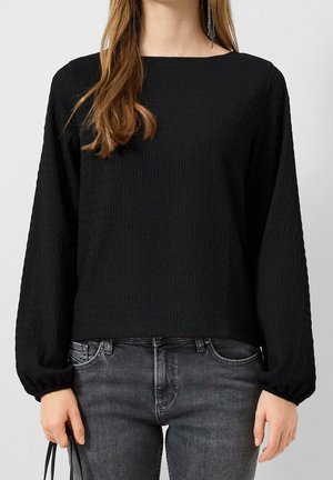 Kvinde iført en tekstureret sort bluse med lange ærmer med puffede manchetter og falmede sorte jeans, der holder en sort rem i venstre hånd.