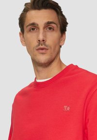 Czerwona bluza z okrągłym dekoltem, z małym wytłoczonym logo po lewej stronie klatki piersiowej oraz teksturowanym materiałem. Prosty design, bez wzorów.