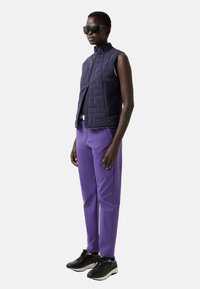 Marineblaue, gesteppte ärmellose Weste mit hohem Kragen, kombiniert mit lila Hosen und schwarzen Turnschuhen. Mit Reißverschluss und Seitentaschen ausgestattet.