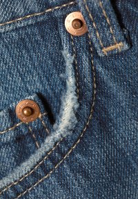 Gros plan sur un tissu en denim bleu avec des bords effilochés, des coutures couleur acajou et des rivets en cuivre sur une section de poche.