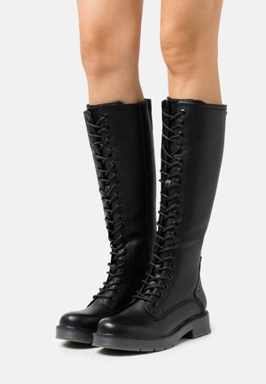 Bottes noires montantes à lacets avec fermeture éclair latérale portées sur des jambes nues, dotées de semelles épaisses en caoutchouc et d'orteils arrondis.