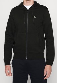 Svart zip-up hoodie i mjukt tyg, med en känguruficka, dragsko på huvan och liten broderad logotyp på vänster bröst.