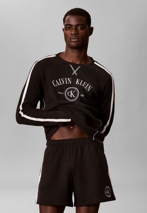 Jonge man die een zwarte Calvin Klein longsleeve en bijpassende korte broek met witte strepen draagt, heft zijn shirt op om zijn buik te laten zien.