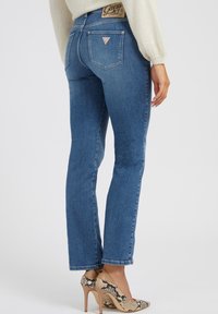 Jeans de mezclilla azul con un corte ajustado, diseño de cinco bolsillos y un pequeño parche con el logo en la parte trasera de la cintura. Combinados con tacones altos con estampado de serpiente.