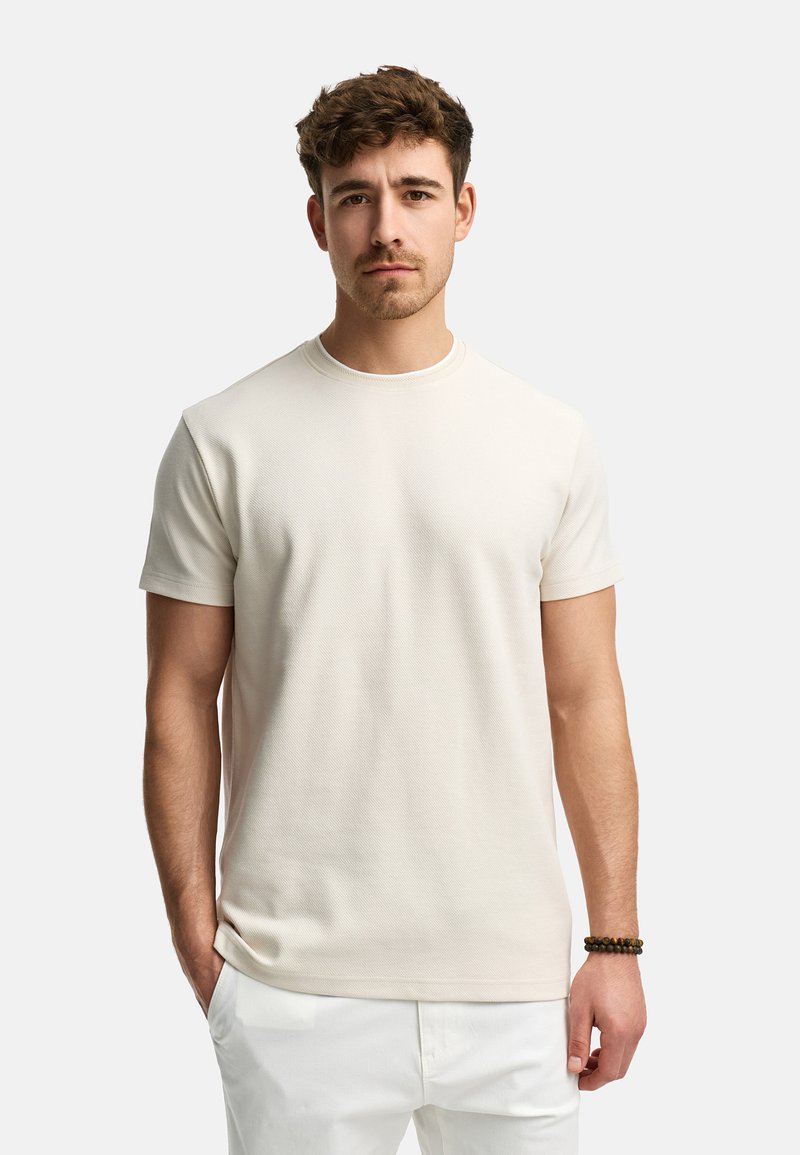 Camiseta de corte corto y de manga corta color beige claro con cuello redondo, fabricada en un tejido texturizado, de forma ajustada y con detalles de costura sutiles. Se lleva con pantalones blancos.