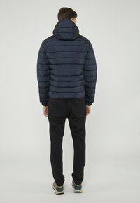 Giacca a vento blu navy con imbottitura orizzontale, cappuccio attaccato e polsini elasticizzati, abbinata a pantaloni neri e scarpe multicolori.