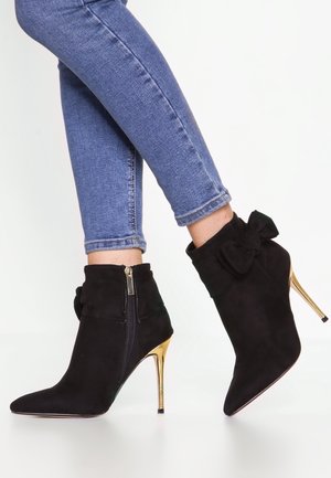 Persoon draagt een blauwe skinny jeans en zwarte suède enkellaarsjes met gouden naaldhakken en decoratieve strikjes aan de zijkant, staand op een witte vloer.