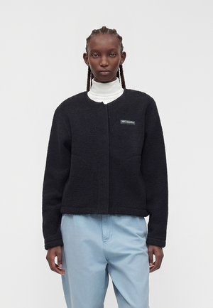Persona con chaqueta de forro polar Columbia negra sobre un jersey de cuello alto blanco y pantalones azul claro, de pie frente a un fondo liso.