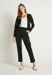 Musta räätälöity puku, jonka kanssa on sovitettu blazer ja kapeat housut, yhdistetty valkoiseen paitaan ja paksuun ruskeaan kaulaketjuun. Valkoiset liukumäkitossut.