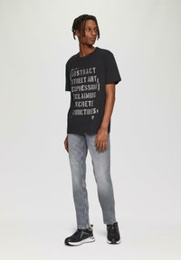 Czarny bawełniany T-shirt z białym napisem graficznym, szare dżinsy oraz czarne sportowe buty z białymi podeszwami, stojący na jednolitym tle.