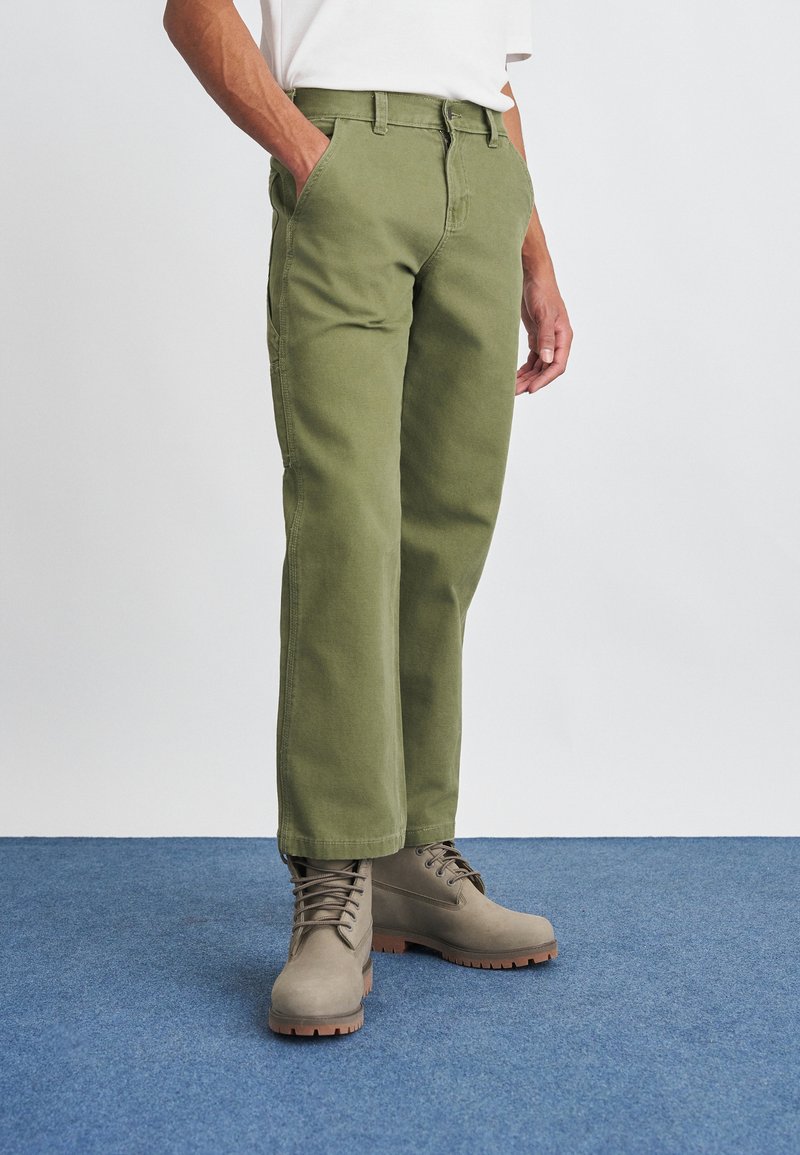 Homem sentado em calças de ganga verde-oliva de perna reta e botas bege com atacadores, de pé sobre um tapete azul com as mãos nos bolsos.