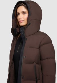 Giacca imbottita marrone con cappuccio, cerniera frontale, bottoni a pressione e dettagli neri. Realizzata con sezioni trapuntate per l'isolamento termico.