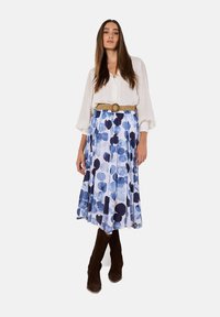 Witte blouse met knopen, blauw-wit patroon, geplooide rok met grote cirkels, beige riem met ronde gesp, en bruine suède laarzen.
