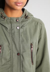 Kvinna som bär en grön huva-jacka med bronserande tryckknappar och vertikala dragkedjefickor på bröstet, läppar och nedre del av ansiktet synliga.