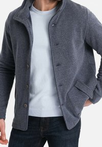 Tmavě šedá bunda vyrobená z měkké tkaniny s límečkem, předním zapínáním na knoflíky a bočními kapsami. Nositá přes světle modrý svetr, kombinovaná s tmavými džíny.