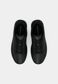 Ună pereche de sneakersi negri Tommy Hilfiger cu șireturi, logo pe lateral și pe limba pantofului, prezentați din sus pe un fundal alb.