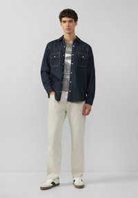 Denim shirt met twee borstzakken, donkerblauw met oranje stiksels, gedragen over een grijze graphic t-shirt. Lichtgekleurde, loszittende broek en sneakers.