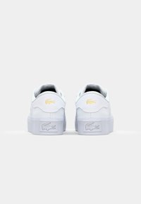 Baskets blanches avec une tige en cuir lisse, des lacets plats et une semelle en caoutchouc texturé. Présente un accent logo doré au niveau du talon.