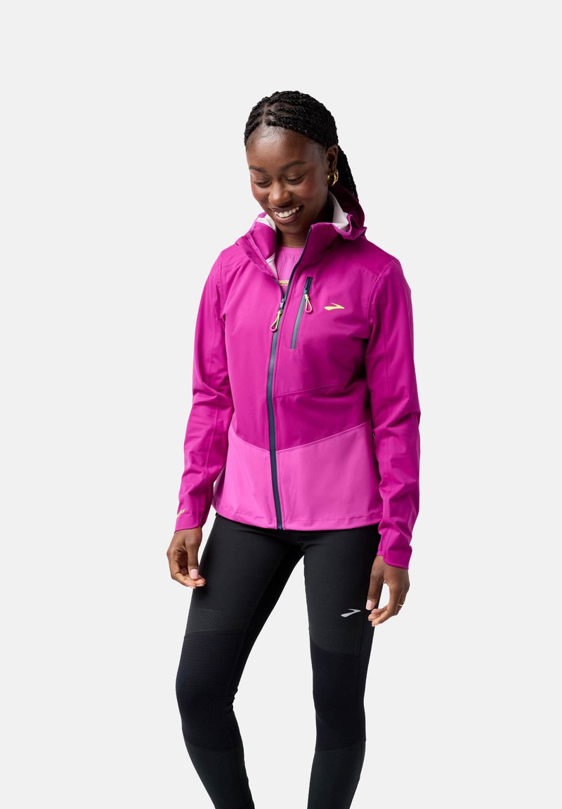 Roze softshell jack met capuchon, ritszakken en contrasterende lichtere secties. Draag het met zwarte leggings met textuurelementen.