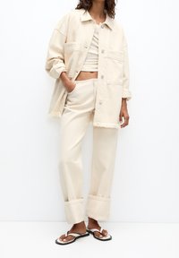 Giacca oversize beige in cotone con due tasche frontali e orlo sfrangiato, abbinata a pantaloni a vita alta coordinati e infradito nere.