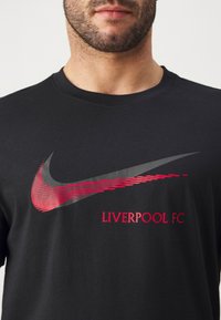 Musta puuvillainen t-paita, jossa on harmaa ja punainen Nike-logo sekä punainen "LIVERPOOL FC" -teksti, lyhyet hihat ja pyöreä pääntie.