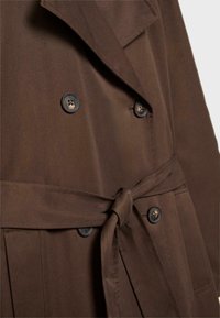 Bruine trenchcoat met een dubbelzijdig ontwerp, grote zwarte knopen, een riem in de taille en een gladde stoftextuur.