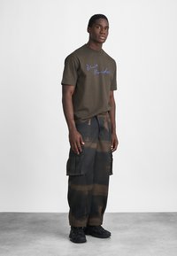 Mann som står iført en brun "Blue Monday" t-skjorte, svarte og brune tie-dye cargo-bukser, og svarte sneakers foran en ensfarget hvit bakgrunn.