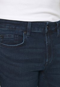 Vaqueros de mezclilla azul oscuro con un corte slim, que cuentan con un bolsillo frontal, cierre de botón y un sutil desgaste en la textura de la tela.