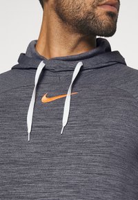 Grau strukturierter Hoodie mit weißem Kordelzug; verfügt über ein orangenes Nike-Logo auf der Brust. Nahaufnahme zeigt das Design des Kragens und der Schulter.