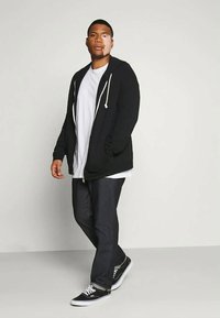 Black zip-up hoodie with white drawstrings over a white t-shirt, paired with dark jeans and black sneakers, standing against a plain background.