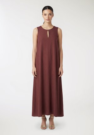 Donna in piedi che indossa un abito senza maniche, lungo fino alla caviglia, di colore marrone rossiccio scuro con una piccola apertura a goccia sul collo e tacchi aperti dorati.