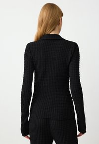 Zwarte gebreide jas met een_TEXTUUR WEAVE, lange mouwen en een afgestikte kraag. Het ontwerp is aansluitend en reikt tot de heupen.