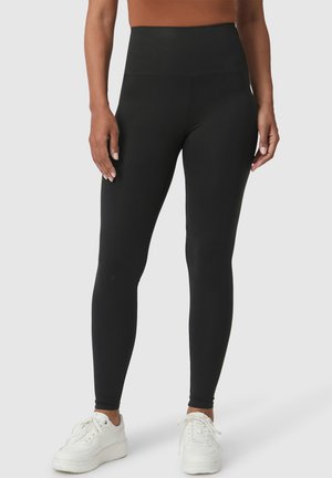 Zwarte high-waisted leggings van rekbaar materiaal, met een gladde afwerking en naadloos ontwerp, gecombineerd met witte sportschoenen.