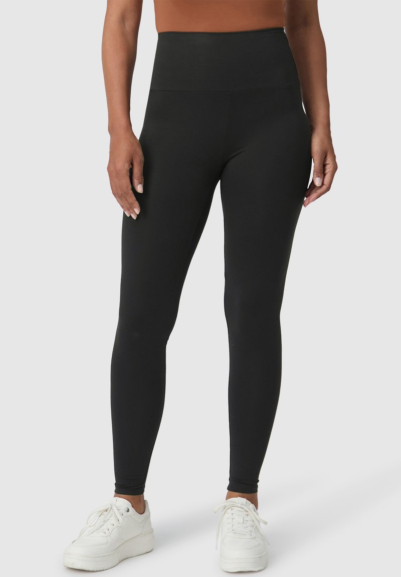 Zwarte high-waisted leggings van rekbaar materiaal, met een gladde afwerking en naadloos ontwerp, gecombineerd met witte sportschoenen.