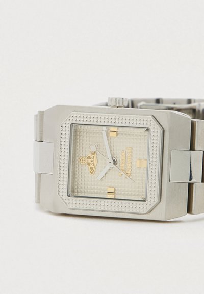 Ασημένιο ορθογώνιο ρολόι χειρός με υφή στο καντράν, χρυσές λεπτομέρειες και μπρασελέ από συνδεδεμένα μεταλλικά στοιχεία που ακουμπάει σε λευκή επιφάνεια.