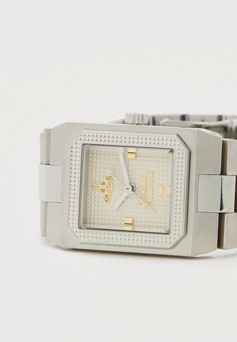 Zilverkleurig rechthoekig polshorloge met een gestructureerde wijzerplaat, gouden accenten en een metalen schakelband, rustend op een wit oppervlak.