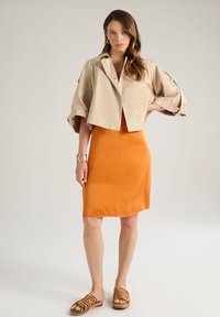 Giacca beige cropped con maniche voluminose, abbinata a un vestito arancione aderente e sandali marroni con cinturini. Bracciale d'oro al polso.
