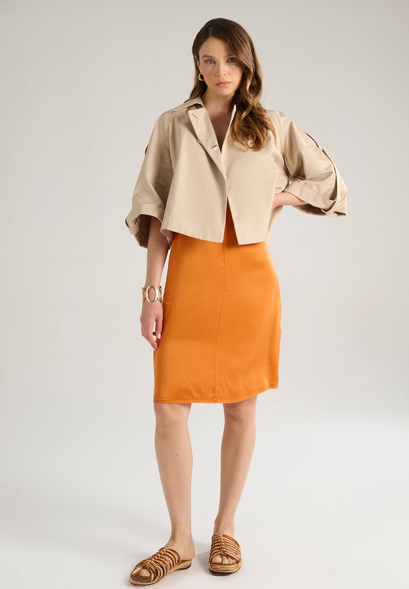 Giacca beige cropped con maniche voluminose, abbinata a un vestito arancione aderente e sandali marroni con cinturini. Bracciale d'oro al polso.