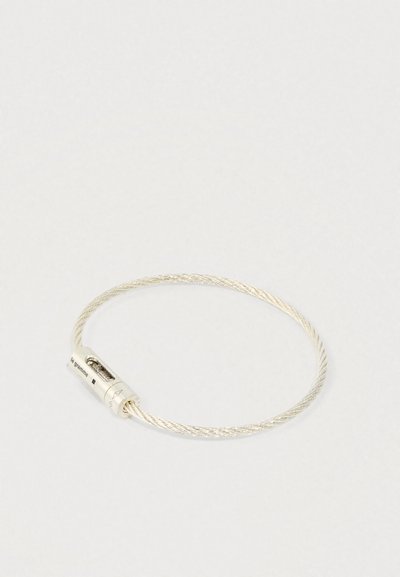 Sudrabota spirāļu vadu aproce ar cilindrisku aizdari, kurā ir iebūvēts ruļļi un mazs gravēts teksts uz vienkārša fona.