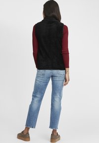 Svart fluffig väst med hög krage, bärs över en vinröd ribbad topp. Ljustvättade raka jeans och bruna rutiga platåskor.