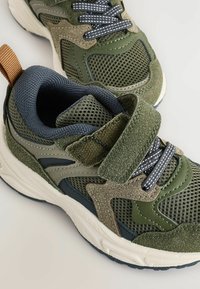Zielone buty sportowe z połączeniem materiałów siateczkowych i zamszowych, z teksturowanymi detalami, regulowanym paskiem i niebieskimi sznurówkami akcentującymi.