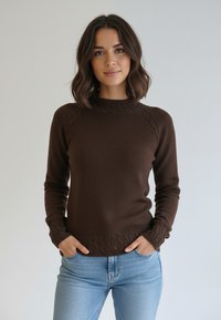 Brązowy sweter dziany z wysokim kołnierzem, z prążkowanymi detalami na ramionach i mankietach oraz fakturowanym wzorem wzdłuż krawędzi. Połączony z niebieskimi dżinsami.