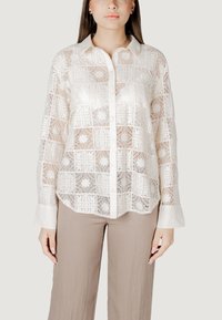 Valge pitsbluus keeruka ruutmustriga, millel on krae, nööpidega esiosa ja pikad varrukad. Kombineeritud beežide laiade pükstega.